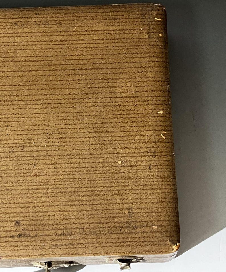 Puede incluir: Una caja rectangular de color marr&oacute;n con una superficie texturizada y acanalada. La caja presenta una esquina visible con un ligero desgaste y peque&ntilde;as rozaduras claras. Un mecanismo de cierre met&aacute;lico es parcialmente visible en el borde.