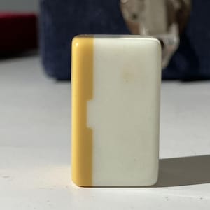 May include: A rectangular mahjong tile standing upright, featuring a smooth, glossy finish. The tile is divided into two vertical sections, with a narrow yellow strip on the left side and a larger, solid white section covering the rest of the surface.