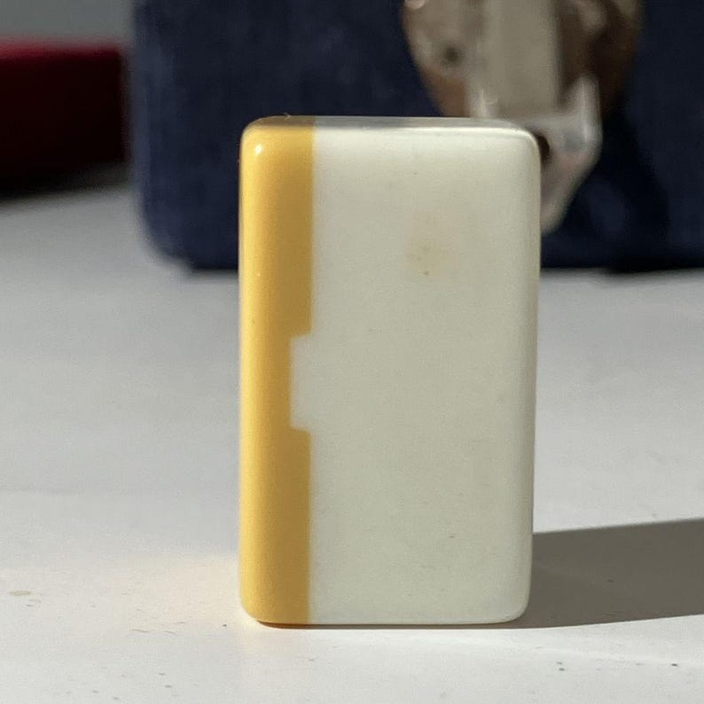 May include: A rectangular mahjong tile standing upright, featuring a smooth, glossy finish. The tile is divided into two vertical sections, with a narrow yellow strip on the left side and a larger, solid white section covering the rest of the surface.