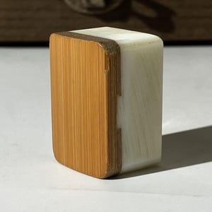 Puede incluir: Un bloque rectangular con un panel frontal de madera de color marr&oacute;n claro con veta vertical, unido a un cuerpo de resina opaca de color blanco roto. El objeto tiene esquinas redondeadas y una construcci&oacute;n s&oacute;lida y compacta con contraste.