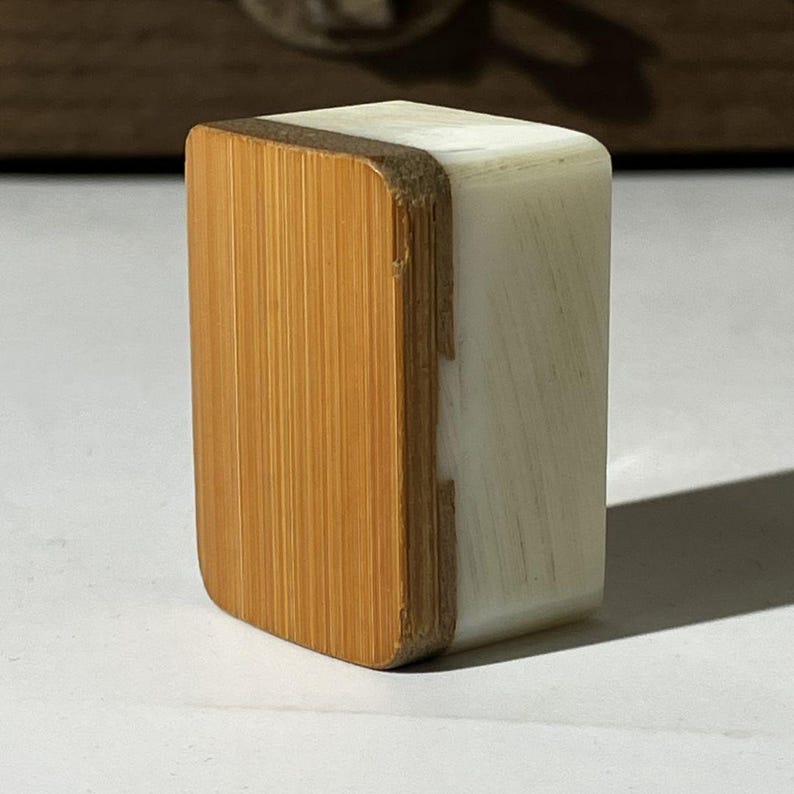 Puede incluir: Un bloque rectangular con un panel frontal de madera de color marr&oacute;n claro con veta vertical, unido a un cuerpo de resina opaca de color blanco roto. El objeto tiene esquinas redondeadas y una construcci&oacute;n s&oacute;lida y compacta con contraste.