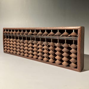 Könnte beinhalten: Ein Vintage-Abakus aus Holz mit einem dunkelbraunen Rahmen und hellbraunen Perlen. Der Abakus hat mehrere Reihen von Perlen, jede mit einer einzigartigen Form. Der Rahmen ist rechteckig und scheint aus massivem Holz zu bestehen.