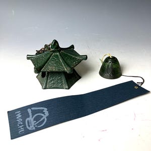 May include: A green metal wind chime with a pagoda-shaped top and a bell hanging from it. The wind chime is on a white surface with a black tag that says "IMACHI" in white lettering.