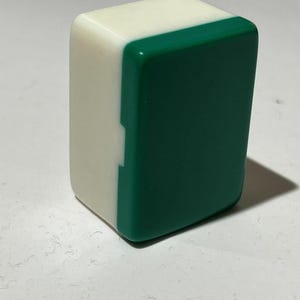 Pu&ograve; includere: Una tessera rettangolare da mahjong posizionata in verticale, caratterizzata da una finitura liscia e lucida. La tessera &egrave; composta da una base solida bianco opaco e da una parte frontale verde scuro brillante, con una piccola tacca sul lato.
