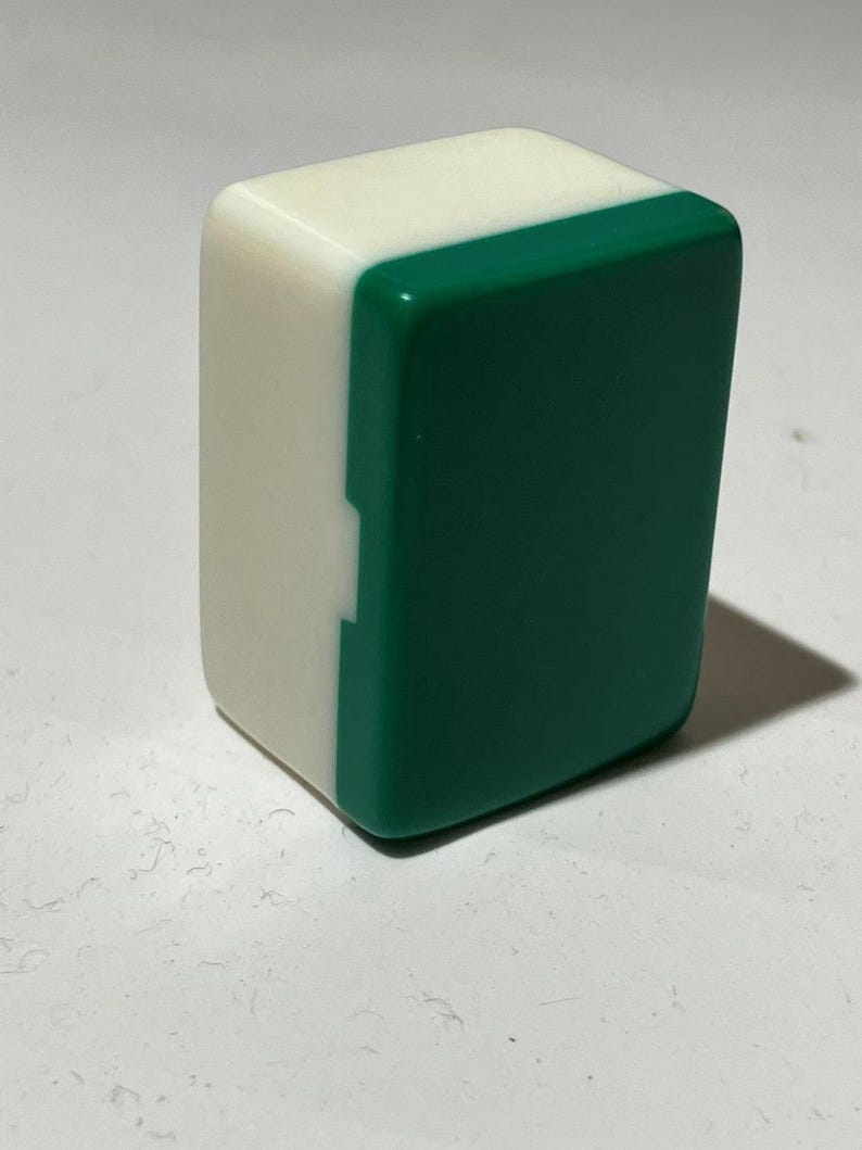Pu&ograve; includere: Una tessera rettangolare da mahjong posizionata in verticale, caratterizzata da una finitura liscia e lucida. La tessera &egrave; composta da una base solida bianco opaco e da una parte frontale verde scuro brillante, con una piccola tacca sul lato.