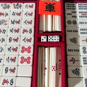 May include: A mahjong set featuring off-white rectangular tiles with black, red, and green engraved characters and symbols. The set includes a bright orange tile with the black character for East, three white dice with black pips, and a red velvet-lined case.