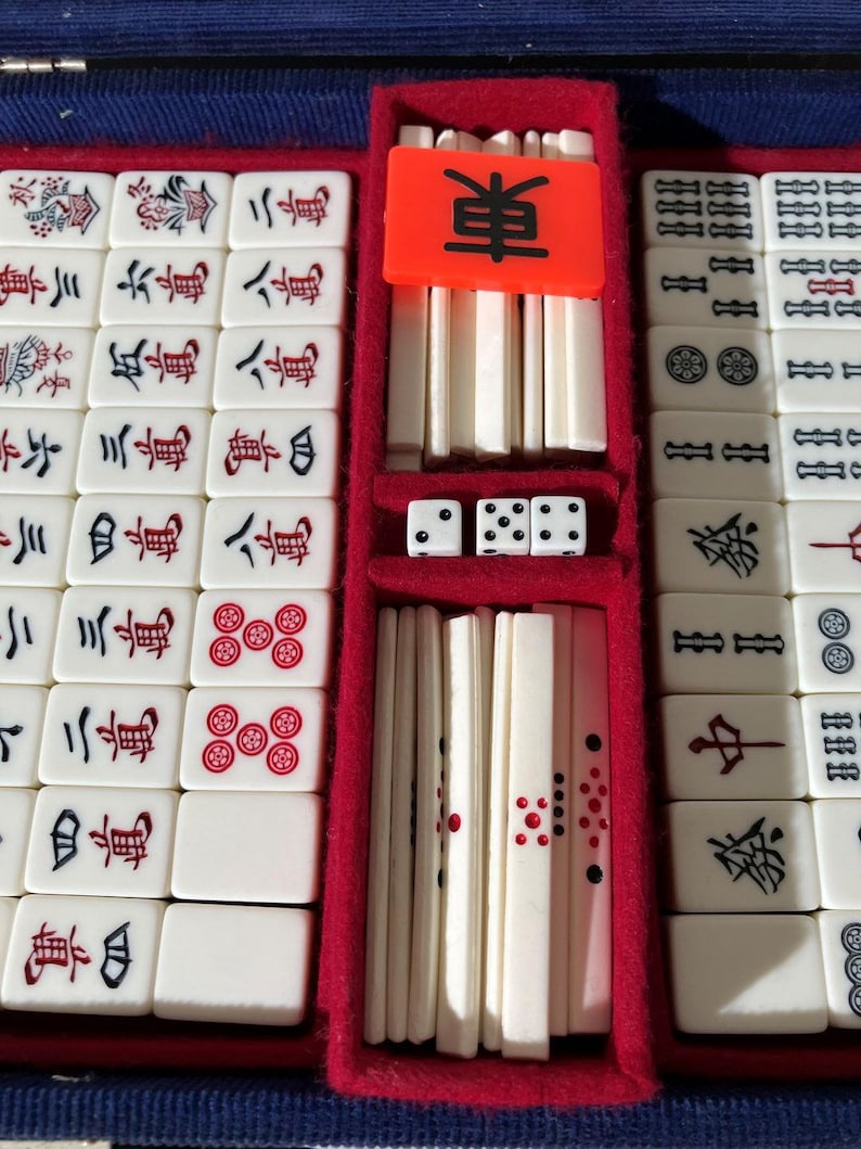 May include: A mahjong set featuring off-white rectangular tiles with black, red, and green engraved characters and symbols. The set includes a bright orange tile with the black character for East, three white dice with black pips, and a red velvet-lined case.