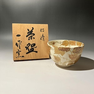 Puede incluir: Un cuenco de cerámica color crema con diseños de hojas y vides en pan de oro. A la izquierda del cuenco hay una placa de madera con caracteres japoneses. El cuenco tiene forma redondeada y superficie lisa.