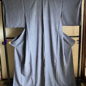 Puede incluir: Un kimono gris cuelga de un perchero de metal negro. La prenda tradicional japonesa está hecha de una tela suave y fluida. El kimono se exhibe en interiores, con una estera de tatami visible debajo.
