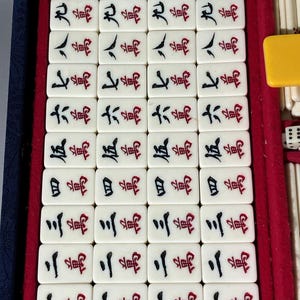 May include: A vintage Mahjong set with ivory-colored tiles arranged in a blue velvet-lined case. The tiles feature Chinese characters in black and red ink. A yellow tile and dice are visible.