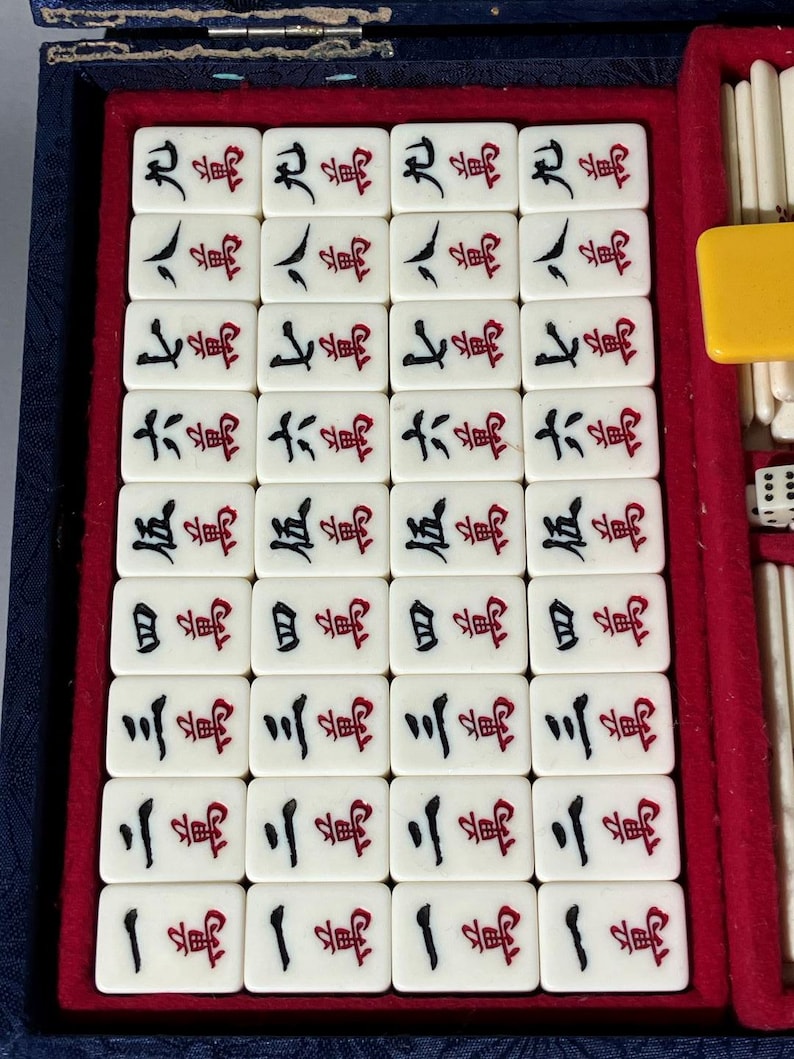 May include: A vintage Mahjong set with ivory-colored tiles arranged in a blue velvet-lined case. The tiles feature Chinese characters in black and red ink. A yellow tile and dice are visible.