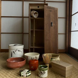 Puede incluir: Una colección de artículos para la ceremonia del té japonesa, que incluye cuencos de cerámica, un batidor de bambú y un armario de madera. Los cuencos varían en tamaño, forma y color, algunos con diseños pintados. El armario es de madera oscura.