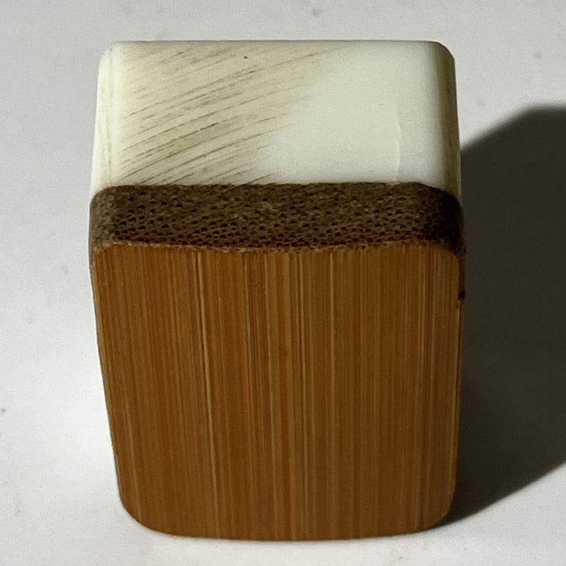 Puede incluir: Una ficha de mahjong rectangular que presenta una secci&oacute;n superior blanca lisa con sutiles estr&iacute;as diagonales y una secci&oacute;n inferior m&aacute;s grande de bamb&uacute; marr&oacute;n claro con veta vertical visible y una banda horizontal texturizada.