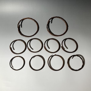 Pode incluir: Uma coleção de dez anéis de metal castanho escuro e desgastado de vários tamanhos. Os anéis estão dispostos em um padrão de grade contra um fundo branco liso. O metal parece ter uma superfície texturizada.