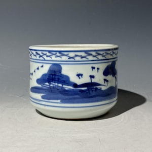 Può includere: Una piccola ciotola rotonda in ceramica bianca con un disegno di paesaggio blu. La ciotola presenta una fascia continua di immagini di paesaggio blu intorno all'esterno, con un bordo blu decorativo sul bordo. Il design include alberi e acqua.