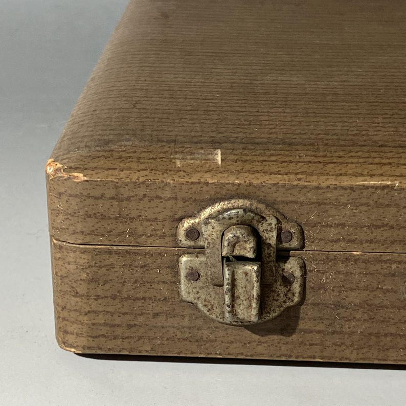 Puede incluir: Una caja de almacenamiento rectangular con un acabado exterior marr&oacute;n texturizado. La caja cuenta con un mecanismo de cierre de metal desgastado centrado en el borde frontal, mostrando signos de oxidaci&oacute;n y desgaste en la superficie.