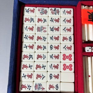 May include: A mahjong set housed in a blue case with a red velvet interior. The set includes off-white tiles featuring black and red Chinese characters and floral motifs, along with white dice and additional game components stored in side compartments.