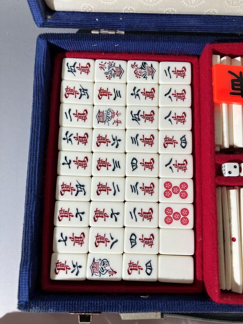 May include: A mahjong set housed in a blue case with a red velvet interior. The set includes off-white tiles featuring black and red Chinese characters and floral motifs, along with white dice and additional game components stored in side compartments.
