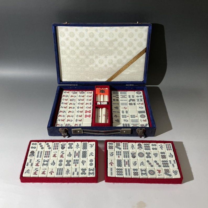 May include: A mahjong set featuring white tiles with black, red, and green characters and symbols. The set includes a blue carrying case with a white patterned interior, two red trays filled with tiles, and a central compartment for counting sticks.