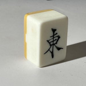 May include: A single rectangular mahjong tile with a white front face featuring the black Chinese character for East. The tile has a smooth, glossy finish and a distinct yellow-toned back section, standing upright to display the engraved character clearly.