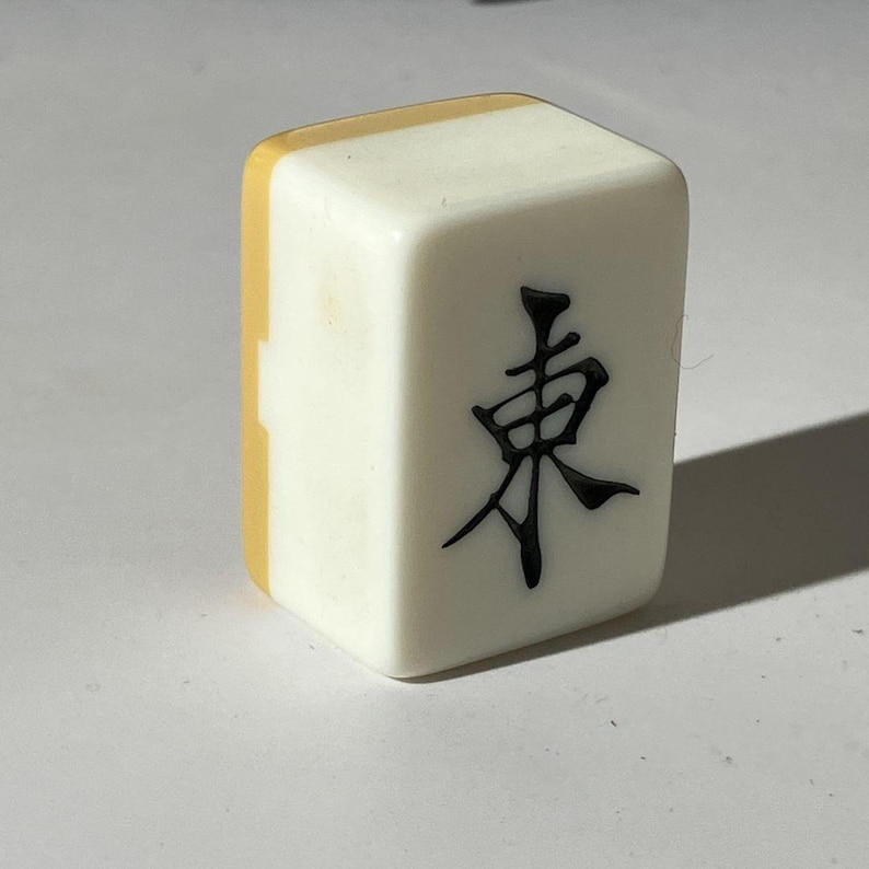 May include: A single rectangular mahjong tile with a white front face featuring the black Chinese character for East. The tile has a smooth, glossy finish and a distinct yellow-toned back section, standing upright to display the engraved character clearly.