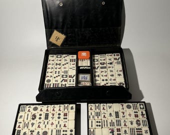 May include: A vintage Mahjong set in a black carrying case, with ivory-colored tiles featuring Chinese characters and symbols. The set includes a variety of tiles, dice, and a small box. Two additional trays of tiles are displayed below the case.