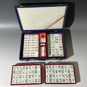 May include: A mahjong set in a blue velvet-lined carrying case with a white patterned interior lid. The set includes numerous white tiles with red, green, and black characters, symbols, and numbers, along with two small white dice and tile racks.
