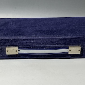 Pu&ograve; includere: Una custodia rettangolare rivestita in tessuto di velluto blu scuro. La custodia presenta una maniglia centrale composta da una striscia di plastica blu e argentata, fissata alle estremit&agrave; da supporti metallici rettangolari con rivetti.