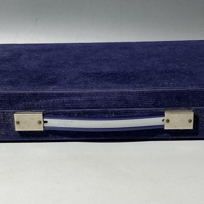 Pu&ograve; includere: Una custodia rettangolare rivestita in tessuto di velluto blu scuro. La custodia presenta una maniglia centrale composta da una striscia di plastica blu e argentata, fissata alle estremit&agrave; da supporti metallici rettangolari con rivetti.