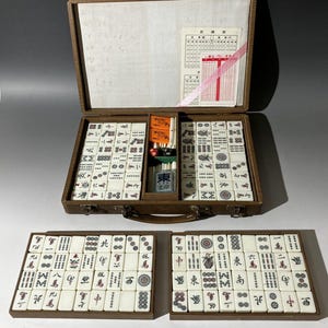 Puede incluir: Un juego de mahjong en un malet&iacute;n marr&oacute;n con una tapa interior blanca que presenta una tarjeta de puntuaci&oacute;n. El conjunto incluye numerosas fichas blancas con s&iacute;mbolos negros, rojos, verdes y azules, junto con marcadores naranjas, dados.