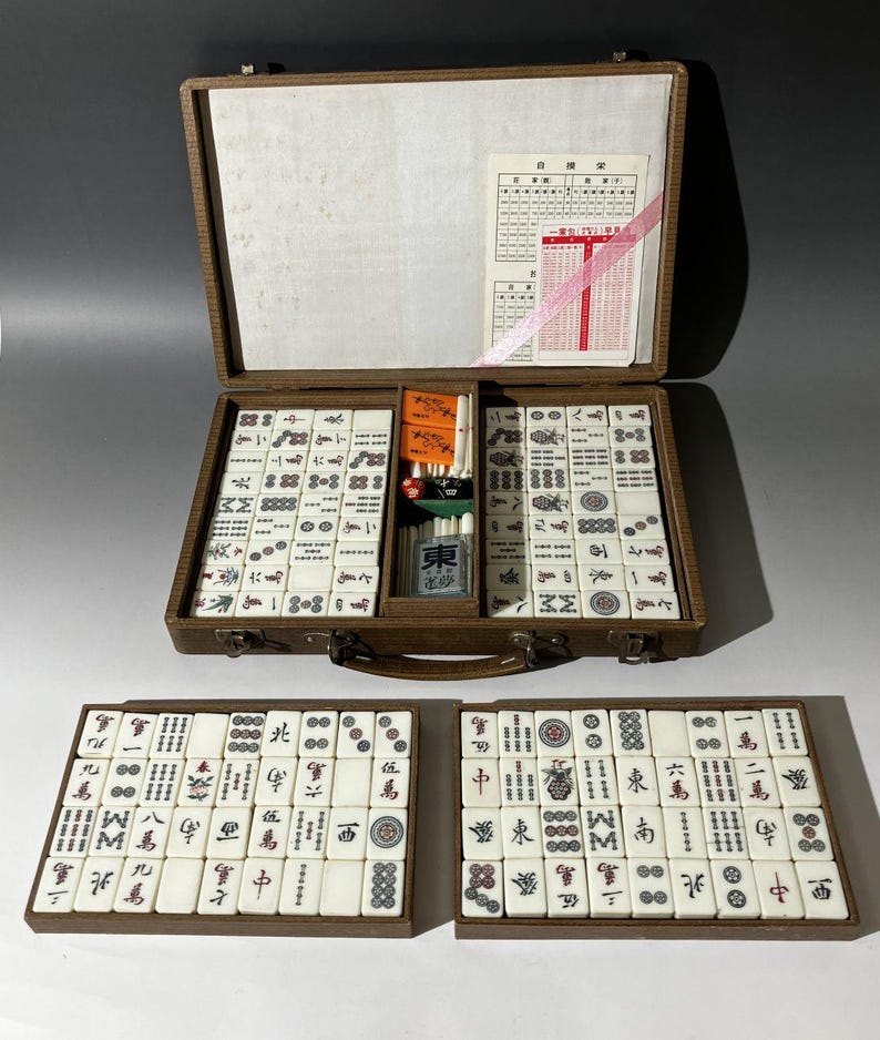 Puede incluir: Un juego de mahjong en un malet&iacute;n marr&oacute;n con una tapa interior blanca que presenta una tarjeta de puntuaci&oacute;n. El conjunto incluye numerosas fichas blancas con s&iacute;mbolos negros, rojos, verdes y azules, junto con marcadores naranjas, dados.
