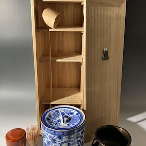 May include: A wooden tea set with a cabinet, a blue and white ceramic container, a small red container, a bamboo whisk, and a black metal bowl. The cabinet has a handle and a wooden cup inside.