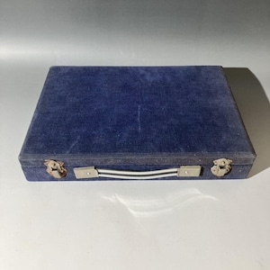 May include: A rectangular storage case covered in dark blue corduroy fabric. The case features a central handle with white and black stripes, secured by silver-toned metal hardware. Two metallic latches are positioned on the front edge.