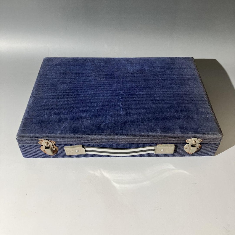 May include: A rectangular storage case covered in dark blue corduroy fabric. The case features a central handle with white and black stripes, secured by silver-toned metal hardware. Two metallic latches are positioned on the front edge.