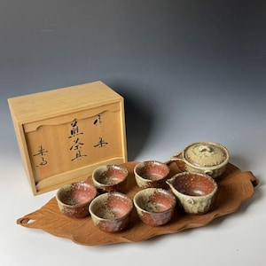 Pode incluir: Um conjunto de chá japonês com uma chaleira, seis xícaras e uma bandeja de madeira. O conjunto é decorado com um esmalte marrom e branco. As xícaras e a chaleira têm um padrão salpicado. A bandeja tem a forma de uma folha. O conjunto está em uma caixa de madeira com escrita japonesa na tampa.