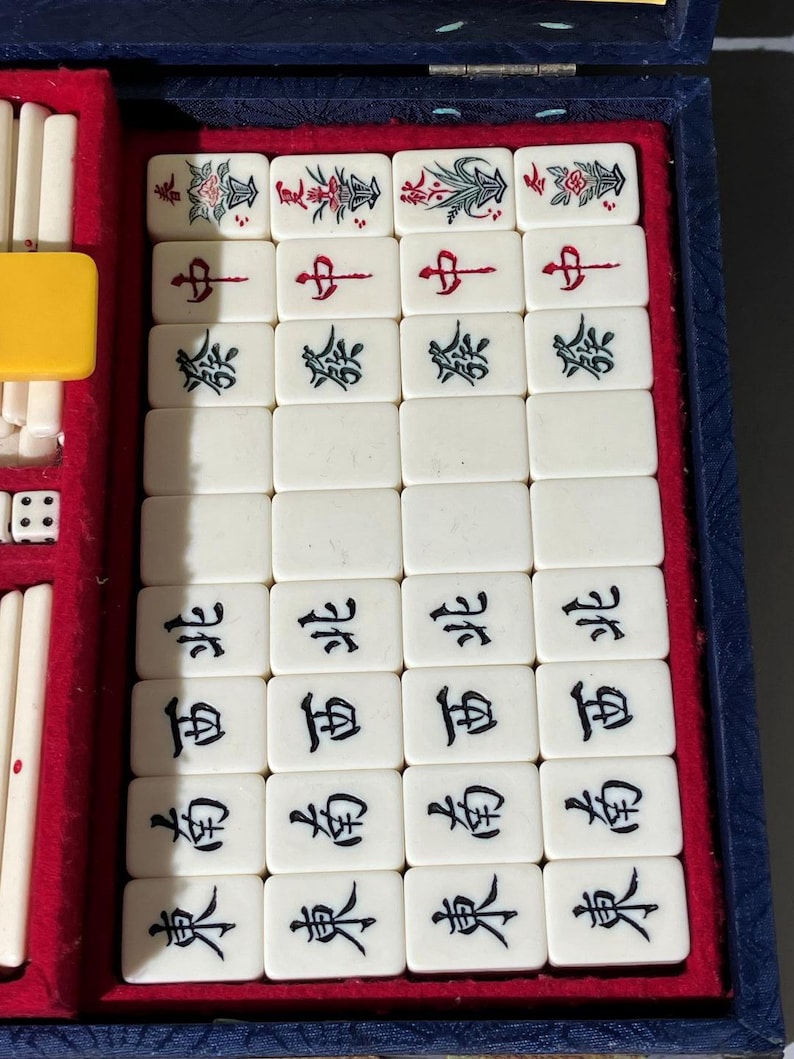 May include: A vintage Mahjong set with ivory-colored tiles, each displaying Chinese characters and decorative floral designs. The set is housed in a blue case with a red velvet interior, including dice and ivory-colored sticks.