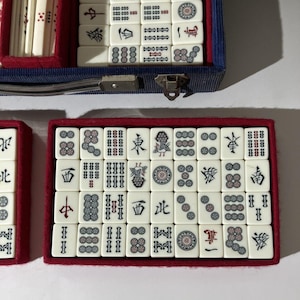 May include: A mahjong set featuring off-white rectangular tiles with black, red, and green engraved symbols, characters, and circular patterns. The tiles are organized in rows within a red-lined blue case with a metal latch and handle.