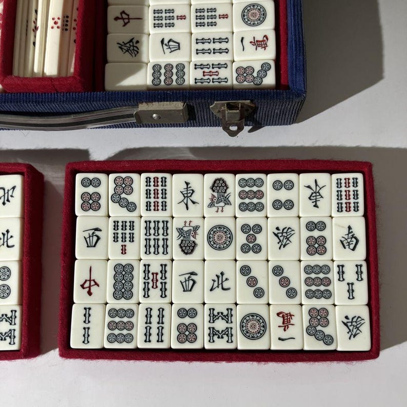 May include: A mahjong set featuring off-white rectangular tiles with black, red, and green engraved symbols, characters, and circular patterns. The tiles are organized in rows within a red-lined blue case with a metal latch and handle.