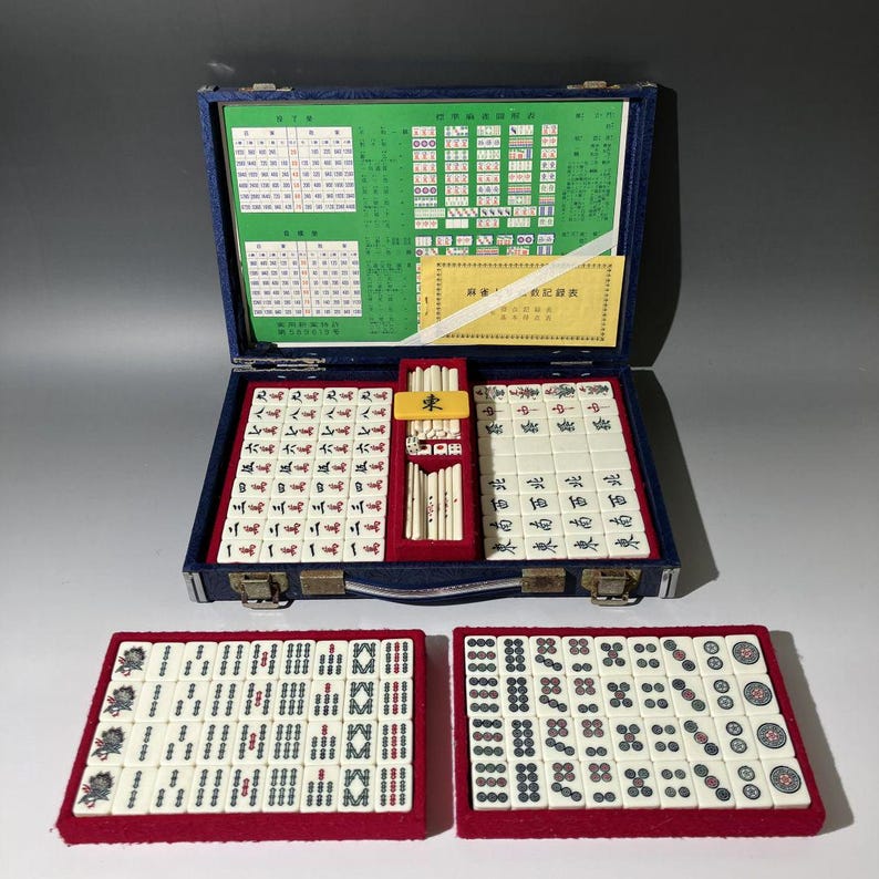 May include: A vintage Mahjong set in a blue velvet case, open to reveal tiles and accessories. Two additional trays of tiles are displayed. The tiles are white with black, red, and green markings. The case has gold-colored clasps.