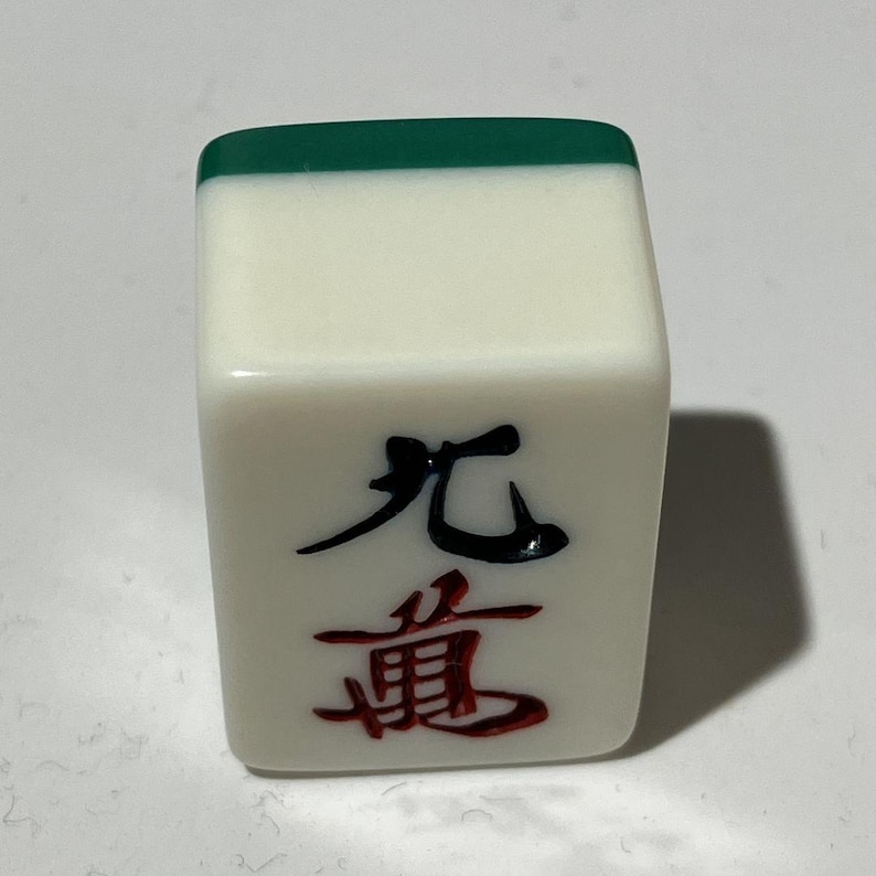Pu&ograve; includere: Una tessera rettangolare da mahjong con una superficie bianca liscia e un bordo superiore verde scuro. La parte anteriore presenta un carattere nero sopra un carattere rosso, entrambi incisi nel materiale. La tessera ha angoli arrotondati.