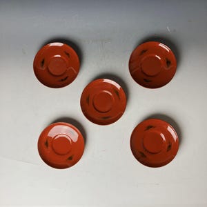 以下が含まれることがあります： 黒い模様の付いた赤い陶器の受け皿が5つ。受け皿は丸く、縁にわずかな盛り上がりがある。