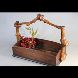 Puede incluir: Una cesta de madera con un asa de rama única. La cesta está hecha de madera oscura y tiene una forma rectangular. El asa está hecha de dos ramas que están conectadas en la parte superior. La cesta está vacía, pero podría usarse para guardar flores, frutas u otros artículos.