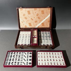 Puede incluir: Un juego de mahjong en un maletín marrón con forro interior estampado. El set incluye fichas blancas con caracteres, símbolos y puntos en negro y rojo, junto con dados blancos pequeños y varillas de conteo en bandejas rojas.