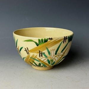 Può includere: Una ciotola in ceramica gialla con un motivo floreale. La ciotola è decorata con fiori bianchi, foglie verdi e dettagli dorati.