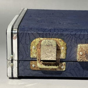 May include: Close-up of a vintage blue case with a textured surface and silver trim. The case features a tarnished metal latch and hinges, showing signs of age and wear. The case's design suggests it may be for musical instruments or tools.