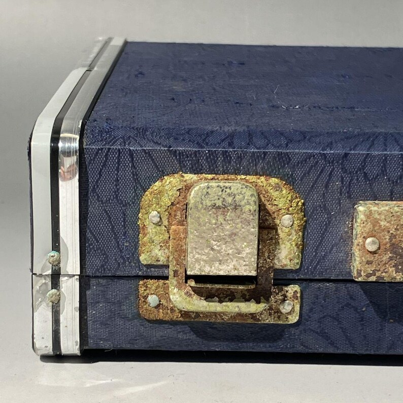 May include: Close-up of a vintage blue case with a textured surface and silver trim. The case features a tarnished metal latch and hinges, showing signs of age and wear. The case's design suggests it may be for musical instruments or tools.