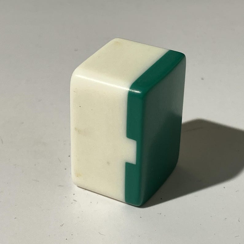 May include: A single, rectangular Mahjong tile. The tile is divided into two sections: a white side and a green side. The green side has a notch cut out of it. The tile is made of a glossy material.