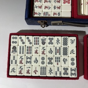 May include: A set of off-white Mahjong tiles with black and red engraved characters and symbols, arranged in a rectangular red-lined tray. The tiles feature various suits, including bamboo, circles, characters, and wind tiles, neatly organized in rows.