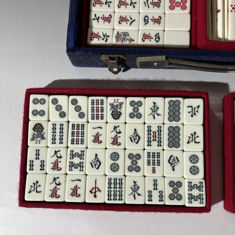 May include: A set of off-white Mahjong tiles with black and red engraved characters and symbols, arranged in a rectangular red-lined tray. The tiles feature various suits, including bamboo, circles, characters, and wind tiles, neatly organized in rows.