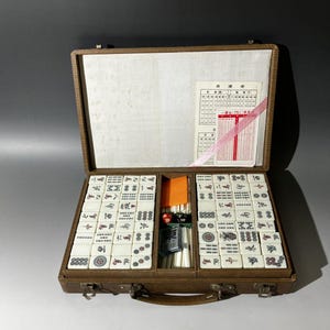 Puede incluir: Un juego de mahjong vintage en un malet&iacute;n marr&oacute;n texturizado con cierres met&aacute;licos. El set incluye fichas blancas con caracteres grabados en negro, rojo y verde, un compartimento central con varillas de conteo blancas, un dado rojo y una tarjeta.